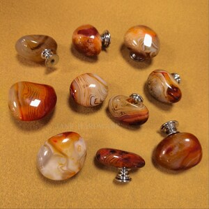 May include: A selection of polished agate cabinet knobs in shades of orange, brown, and cream. Each knob has a unique pattern and is mounted on a silver-toned base. The knobs vary in shape and size, offering a natural, organic aesthetic.