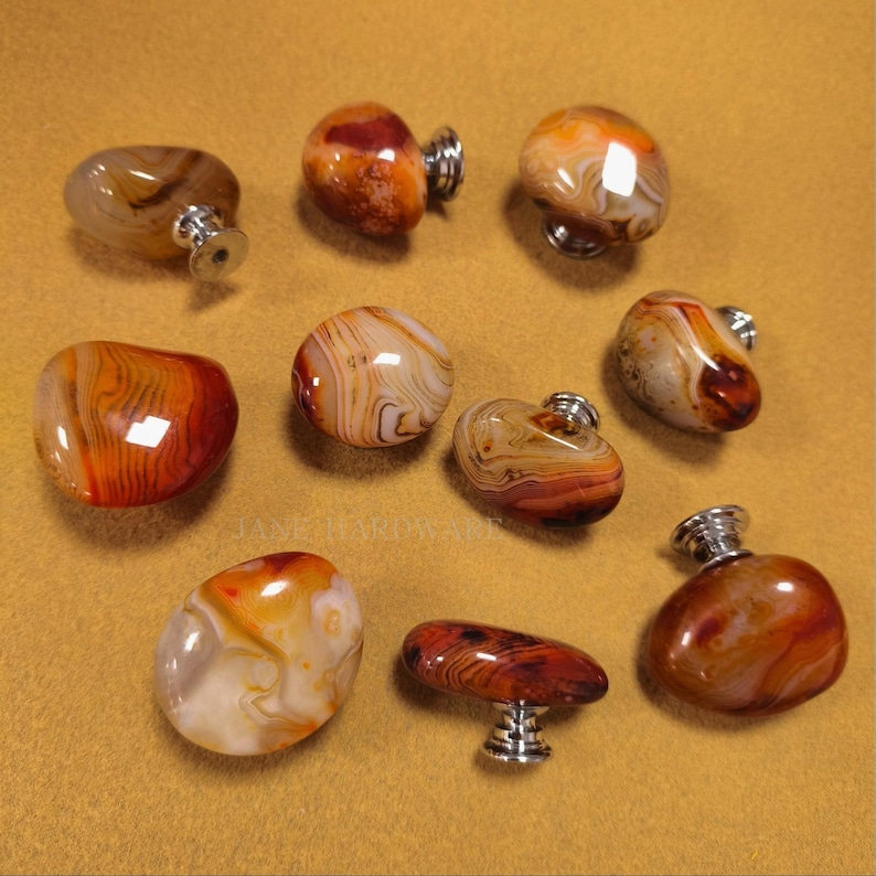 May include: A selection of polished agate cabinet knobs in shades of orange, brown, and cream. Each knob has a unique pattern and is mounted on a silver-toned base. The knobs vary in shape and size, offering a natural, organic aesthetic.