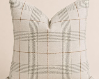 May include: A square throw pillow featuring a light beige fabric base with a woven plaid pattern. The design consists of intersecting thin lines in soft grey and muted brown, creating a grid of squares across the entire surface of the pillow.
