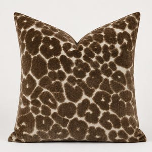 May include: A square throw pillow featuring a textured, fuzzy surface with a dark brown and cream leopard print pattern. The design consists of irregular, rounded dark brown spots set against a lighter cream-colored background fabric.