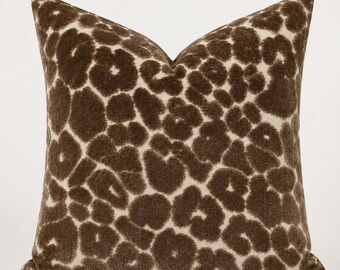 May include: A square throw pillow featuring a textured, fuzzy surface with a dark brown and cream leopard print pattern. The design consists of irregular, rounded dark brown spots set against a lighter cream-colored background fabric.