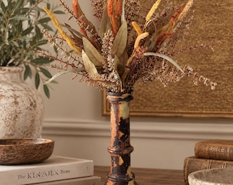 May include: A tall, slender, weathered dark brown and cream-colored pillar-shaped vase holding a dried floral arrangement with orange, yellow, and brown stems, leaves, and grasses. The vase has a textured, distressed finish with visible vertical ridges.
