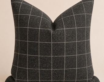May include: A square throw pillow featuring a dark charcoal gray fabric with a subtle, thin white grid pattern. The pillow has a distinct, sharp crease at the top center, creating a structured, plump appearance with clean, straight edges.