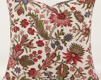 May include: A square throw pillow featuring a cream-colored fabric base with a dense, repeating botanical pattern. The design includes stylized flowers and leaves in shades of deep red, muted olive green, and soft blue, creating a textured appearance.