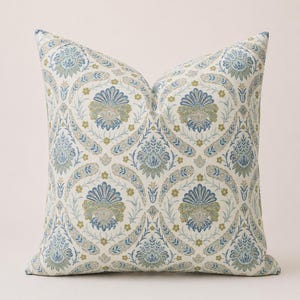 May include: Square decorative pillow with a floral pattern in shades of blue, green, and beige. The design features stylized flowers and paisley-like elements, creating a classic, elegant look. The pillow is likely made of fabric.