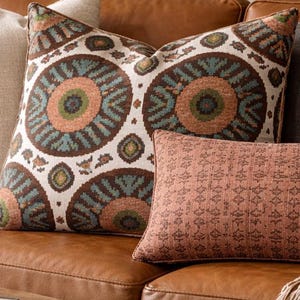 May include: A brown leather sofa with several decorative pillows. The pillows include a patterned pillow with teal, orange, and brown circles, a rectangular pillow with a geometric pattern, and a gray pillow. A beige throw blanket with fringe is draped over the sofa.