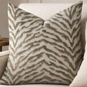 May include: A decorative throw pillow with a zebra stripe pattern in shades of beige and olive green. The pillow is placed on a cream-colored sofa, creating a neutral and stylish aesthetic. The pillow is square-shaped and adds a touch of texture to the room.