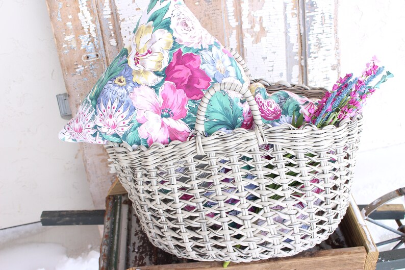 Puede incluir: Una cesta de mimbre blanca con un forro de tela con estampado floral y una almohada floral en su interior. La cesta tiene un asa y est&aacute; llena de ramitas de lavanda.