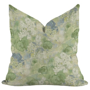 May include: A decorative square pillow with a floral pattern. The pillow features a watercolor design with white, green, and blue hydrangea blooms. The background has a light green and beige linen texture.