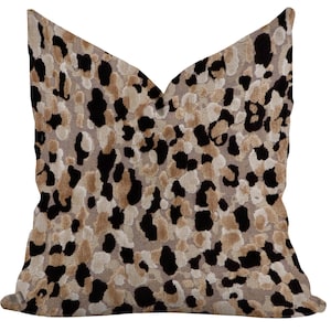 May include: A decorative throw pillow with a leopard print pattern. The pillow features a beige background with black, brown, and cream-colored spots. The pillow has a V-shaped design.