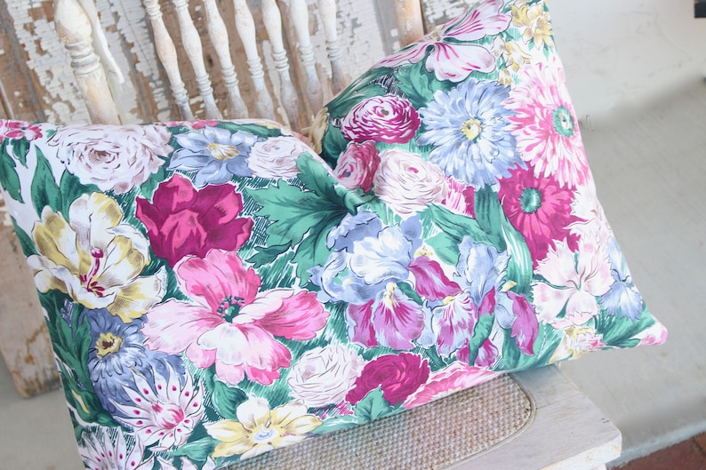 Puede incluir: Dos almohadas decorativas con estampado floral con una variedad de flores rosas, amarillas, azules y verdes sobre un fondo blanco.
