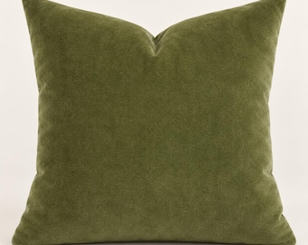 May include: A square throw pillow featuring a solid olive green velvet fabric cover. The pillow has a soft, matte texture with a visible seam along the edges, creating a structured shape with pointed corners and a slight indentation at the top center.