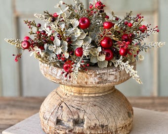 May include: A decorative arrangement of artificial red berries and frosted green foliage sits inside a weathered, round wooden pedestal bowl. The bowl features a distressed, light-colored finish with visible wood grain and natural cracks.