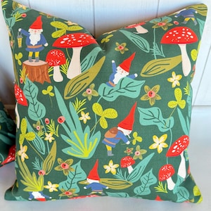 May include: A green pillow with a repeating pattern of gnomes, mushrooms, and leaves. The gnomes are wearing red hats and blue clothing. The mushrooms are red with white stems. The leaves are green and yellow.
