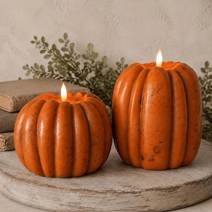 May include: Two orange pumpkin-shaped candles with vertical ridges and a speckled texture. Each candle features a small, lit LED flame at the top center, providing a warm glow. The candles are positioned side-by-side on a circular wooden surface.