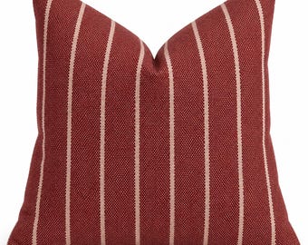 May include: A square throw pillow featuring a deep red, textured fabric base with vertical, thin, light beige stripes running across the front. The pillow has a distinct, plumped center crease and a structured, clean-edged square shape.