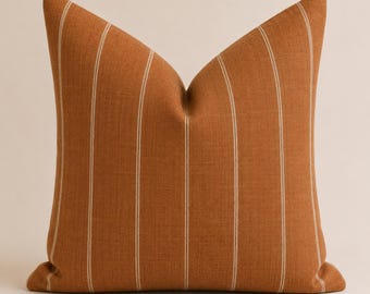 May include: A square throw pillow in a solid burnt orange color featuring vertical pinstripes in a lighter cream tone. The fabric has a visible woven texture, and the pillow is plumped to create a distinct peak at the top center.