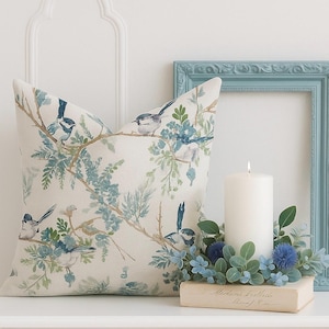 May include: A decorative square pillow with a bird and floral design in shades of blue, green, and beige. The pillow is placed on a white surface next to a lit white candle and a blue picture frame.