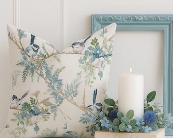 Puede incluir: Un cojín cuadrado decorativo con un diseño de pájaros y flores en tonos azules, verdes y beige. Una vela blanca encendida y un marco de fotos azul se encuentran sobre una superficie blanca.