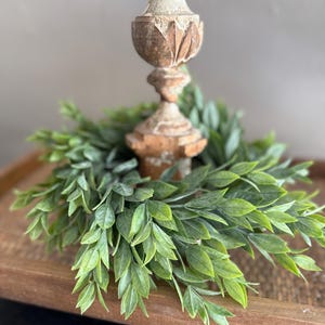 May include: A decorative, distressed wooden candlestick with a green leafy wreath base. The candlestick has a detailed, carved design and a light, neutral color. The wreath is composed of small, vibrant green leaves, creating a natural, rustic aesthetic.