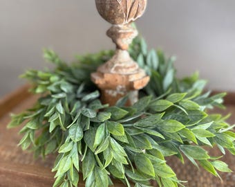 Könnte beinhalten: Ein dekorativer, verwitterter Holzkerzenhalter mit einem grünen Blätterkranz als Basis. Der Kerzenhalter hat ein detailliertes, geschnitztes Design und eine helle, neutrale Farbe. Der Kranz besteht aus kleinen, leuchtend grünen Blättern und erzeugt eine natürliche, rustikale Ästhetik.