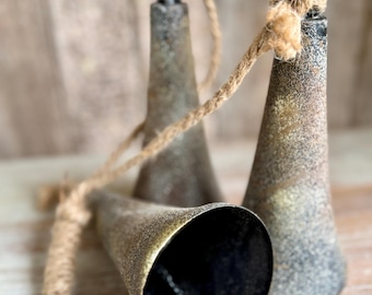 Op de afbeelding: Drie rustieke metalen bellen met een verweerde afwerking en touwhangers. De bellen hebben een donker interieur en een verweerde, getextureerde buitenkant in tinten grijs en goud. Het touw heeft een natuurlijke, bruine kleur.