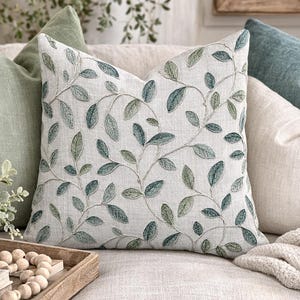 May include: A decorative throw pillow with a botanical leaf design in shades of green and blue on a cream background. The pillow is surrounded by other pillows in shades of green, blue, and cream, creating a cozy and inviting atmosphere.