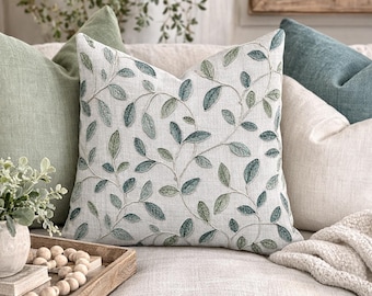 May include: A decorative throw pillow with a botanical leaf design in shades of green and blue on a cream background. The pillow is surrounded by other pillows in shades of green, blue, and cream, creating a cozy and inviting atmosphere.