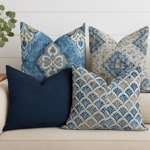 May include: Four decorative throw pillows in shades of blue and beige rest on a cream-colored sofa. The pillows feature various patterns, including a ribbed texture, floral designs, and geometric shapes. The pillows add a touch of elegance to the living space.