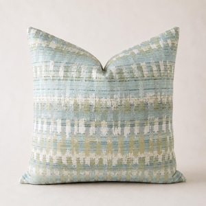 May include: A square decorative pillow with a textured pattern. The pillow features a light blue, green, and white woven design. The pattern consists of vertical and horizontal lines, creating a grid-like appearance. The pillow is a soft, neutral accent for any room.