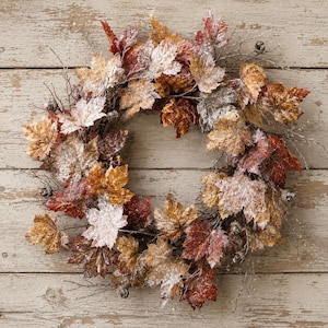 May include: A circular wreath composed of artificial maple leaves in shades of brown, orange, and deep red. The foliage features a light dusting of white frost and is interspersed with small brown acorns and thin, bare, twig-like branches.