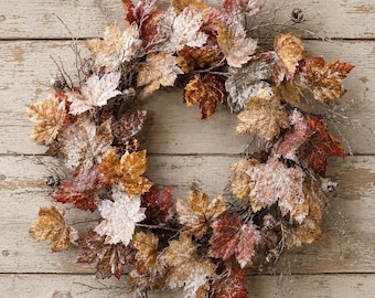 May include: A circular wreath composed of artificial maple leaves in shades of brown, orange, and deep red. The foliage features a light dusting of white frost and is interspersed with small brown acorns and thin, bare, twig-like branches.