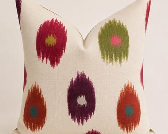 May include: A square throw pillow with an off-white base featuring a repeating pattern of oval-shaped ikat-style motifs. The motifs are colored in shades of magenta, olive green, deep purple, and burnt orange, each with a contrasting center dot.