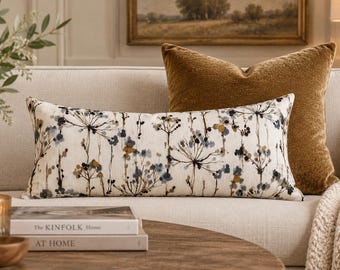 May include: A rectangular lumbar pillow featuring a cream-colored fabric with a botanical print of dark blue, grey, and mustard yellow wildflowers. The pillow is positioned in front of a large, solid, textured mustard yellow square throw pillow.