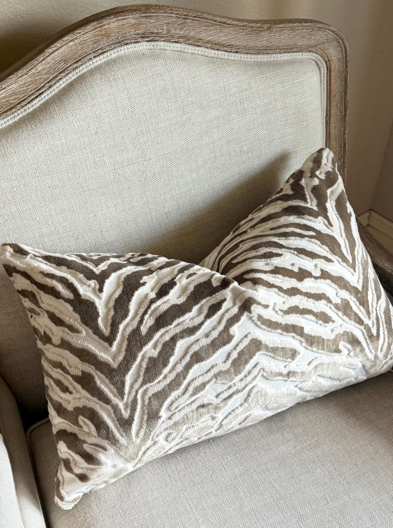 May include: Two decorative throw pillows with a brown and white zebra print pattern. The pillows are on a light beige armchair.