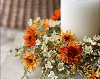 May include: A white pillar candle sits inside a circular floral wreath featuring artificial orange and yellow daisy-like flowers, small white blossoms, and green foliage. The arrangement is constructed on a textured, weathered gray base.