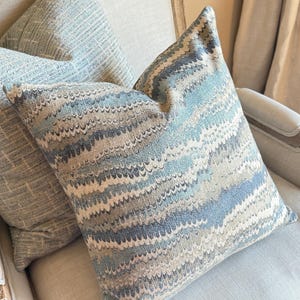 May include: Two decorative throw pillows with a blue and brown wavy pattern. The pillows are on a light beige armchair.