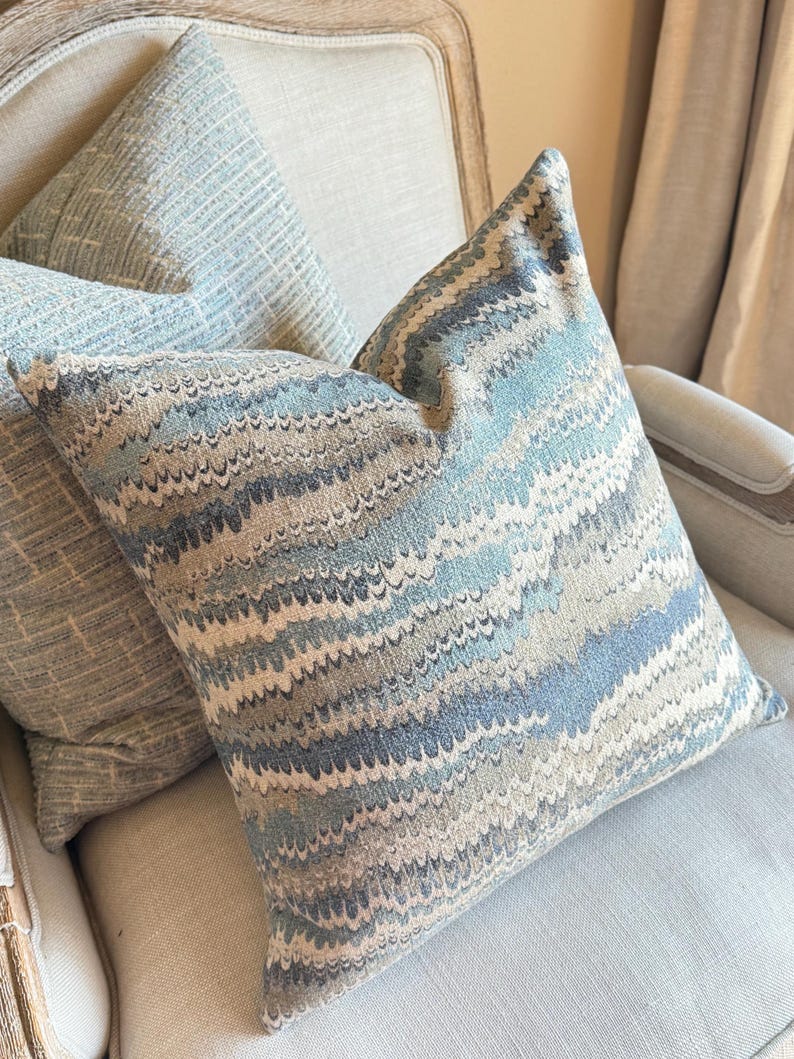 May include: Two decorative throw pillows with a blue and brown wavy pattern. The pillows are on a light beige armchair.