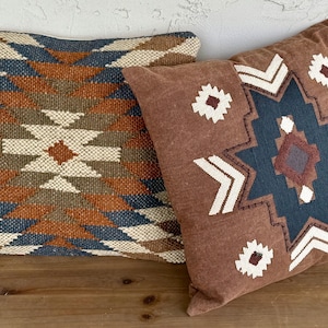 Op de afbeelding: Twee decoratieve sierkussens met geometrische patronen. Eén kussen heeft een bruin, crème en blauw ruitpatroon. Het andere kussen is bruin met een blauw en wit sterpatroon en witte accenten. Beide kussens liggen op een houten oppervlak.