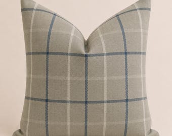 May include: A square throw pillow featuring a light beige base color with a grid pattern composed of thin, intersecting dark blue and off-white lines. The fabric has a visible woven texture, and the pillow is plumped to form a distinct peak.