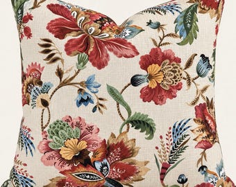 May include: A square throw pillow featuring a cream-colored fabric base with a vibrant floral pattern. The design includes large, stylized blossoms in shades of deep red, golden yellow, and muted blue, accented by intricate green foliage and stems.