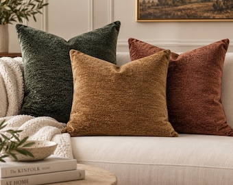 May include: Three square throw pillows in dark green, golden yellow, and burnt orange. Each pillow features a textured, woven fabric finish. The pillows are arranged in a row, resting against a light-colored sofa, showcasing their distinct colors.