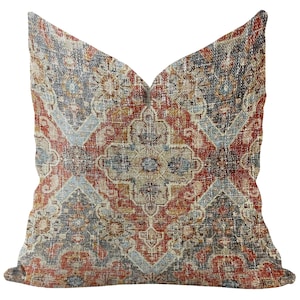 Puede incluir: Cojín cuadrado con un patrón desgastado de estilo vintage. El cojín presenta un diseño de medallón central en tonos de rojo, azul, beige y dorado. La estética general recuerda a una alfombra tradicional.