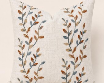 May include: A square throw pillow with a light beige, textured fabric base. The front features two vertical, symmetrical embroidered vine patterns with leaves in shades of rust orange, muted blue, and tan, creating a balanced botanical design.