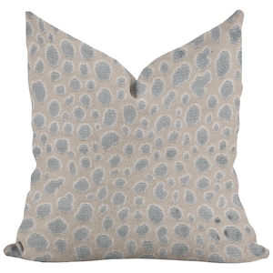 May include: A square decorative pillow with a beige background and a pattern of light blue, irregular-shaped spots. The pillow has a soft, textured appearance, suitable for home decor.