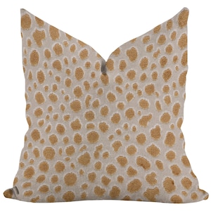 May include: A square decorative pillow with a beige background and a pattern of tan-colored spots. The pillow has a soft, textured appearance, and the spots vary in size and density, creating a spotted design. The pillow is likely used for home decor.
