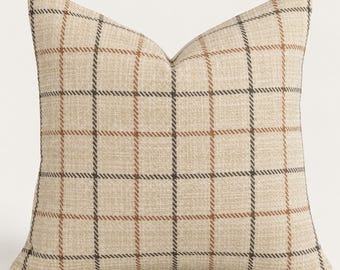May include: A square throw pillow featuring a beige fabric base with a prominent plaid pattern. The design consists of intersecting vertical and horizontal lines in dark charcoal gray and warm brown, creating a classic grid effect across the surface.