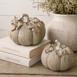 May include: Two decorative pumpkins with a weathered, light brown finish and sculpted leaves around the stems. One pumpkin rests on a stack of two books with the text LIVE BEAUTIFULLY and THIS IS HOME printed on the spines, while the other sits nearby.