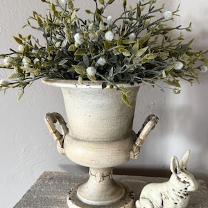 May include: A cream-colored, aged-look urn filled with green and white faux foliage. A small, white rabbit figurine sits beside the urn. The urn has handles and a pedestal base, suggesting a vintage or farmhouse aesthetic.