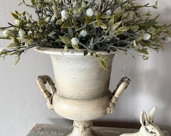 May include: A cream-colored, aged-look urn filled with green and white faux foliage. A small, white rabbit figurine sits beside the urn. The urn has handles and a pedestal base, suggesting a vintage or farmhouse aesthetic.